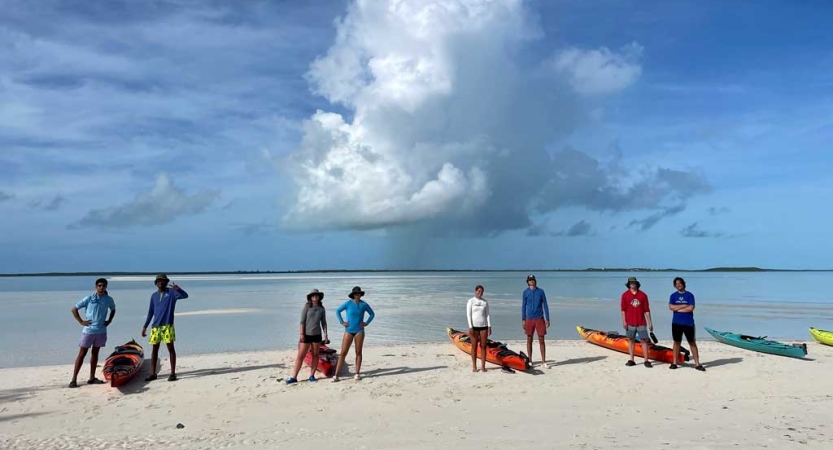 People stand in pairs beside kayaks resting on the beach. 